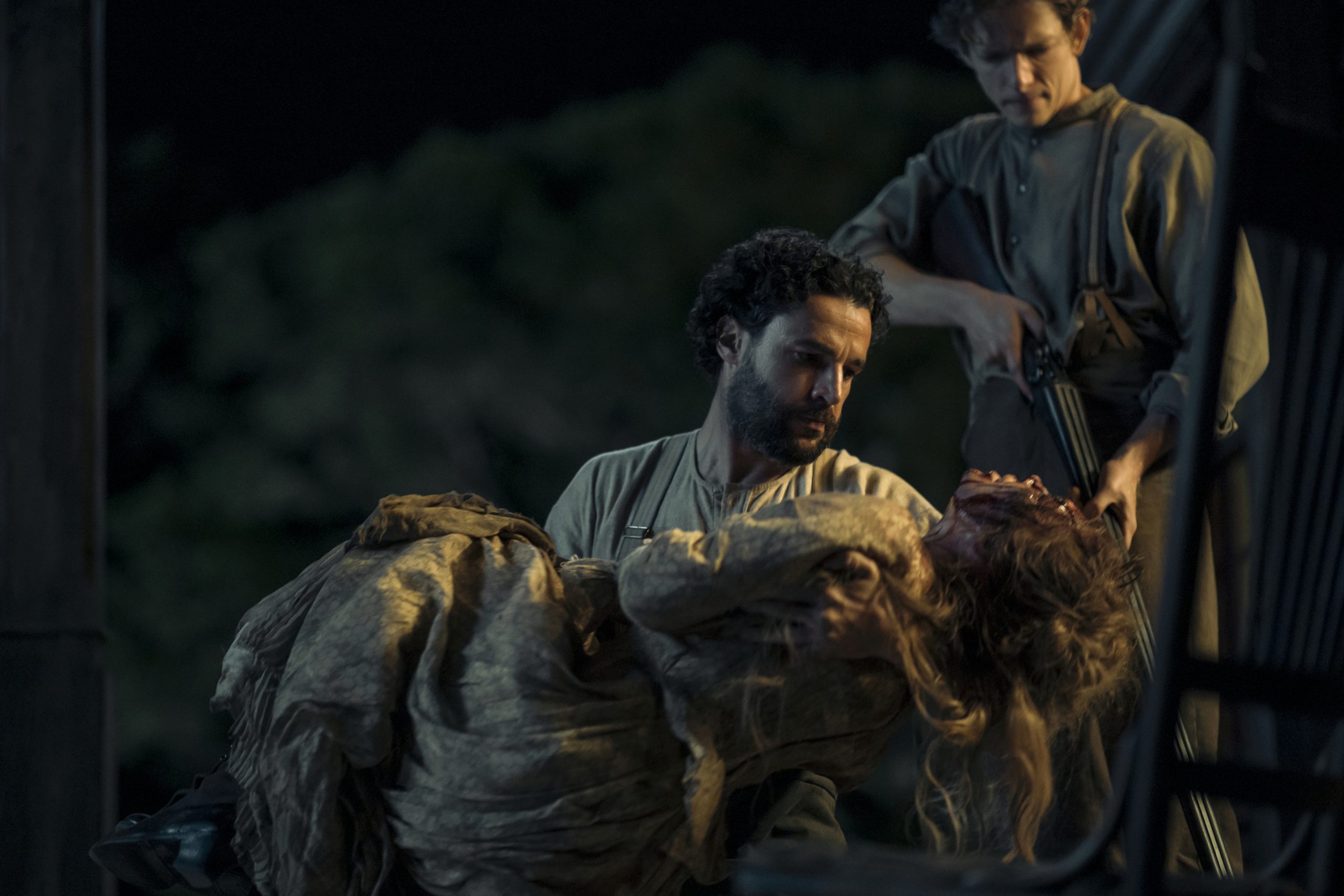 "A man holds an unconscious or injured woman in his arms at night, while another man with a rifle stands behind them outside, suggesting a dramatic or intense situation in a rural or historical setting."