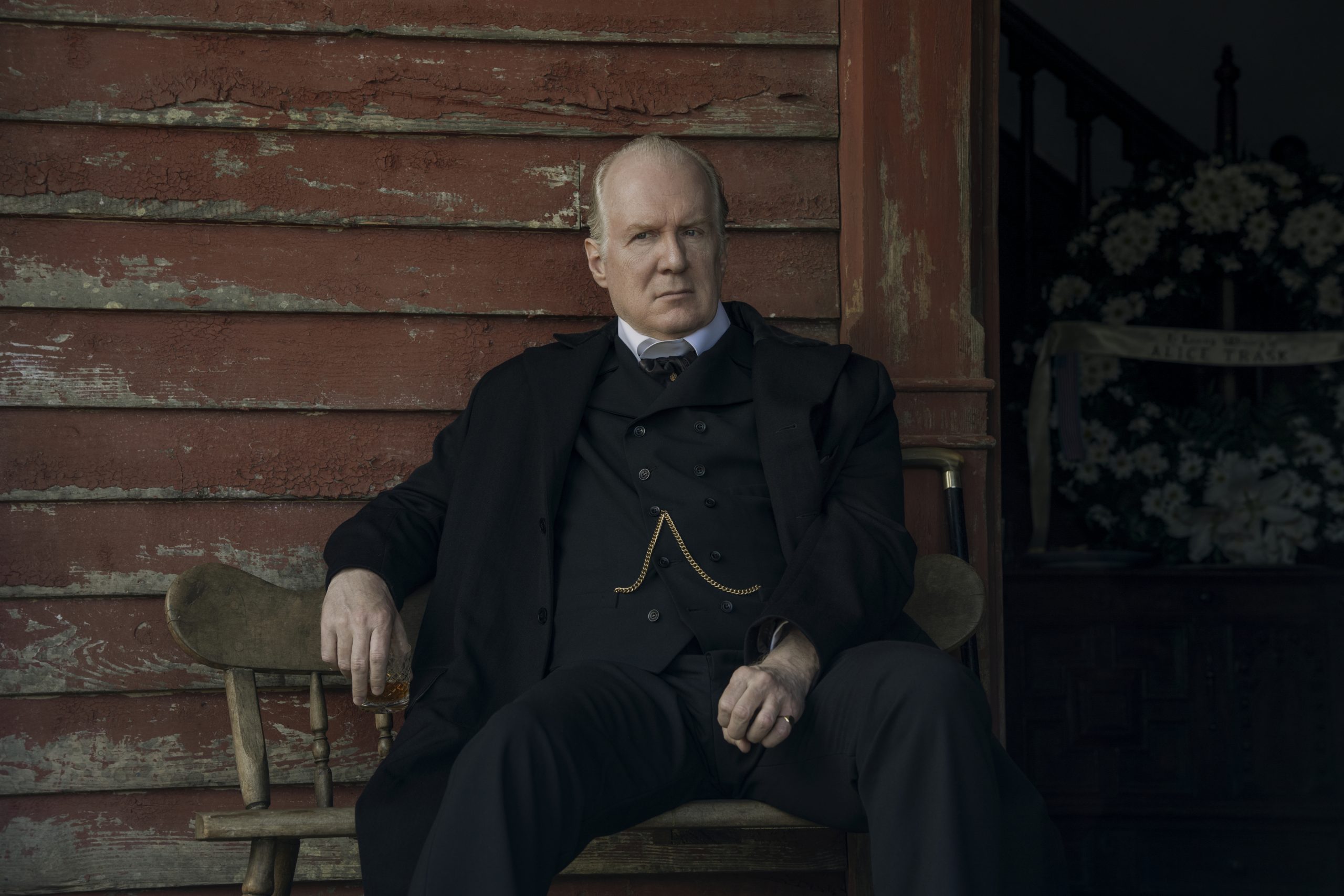 "Stern man in formal black suit sits on bench outside weathered red wooden building, holding a drink; flowers and dark staircase are visible in the background, suggesting a somber or formal event."