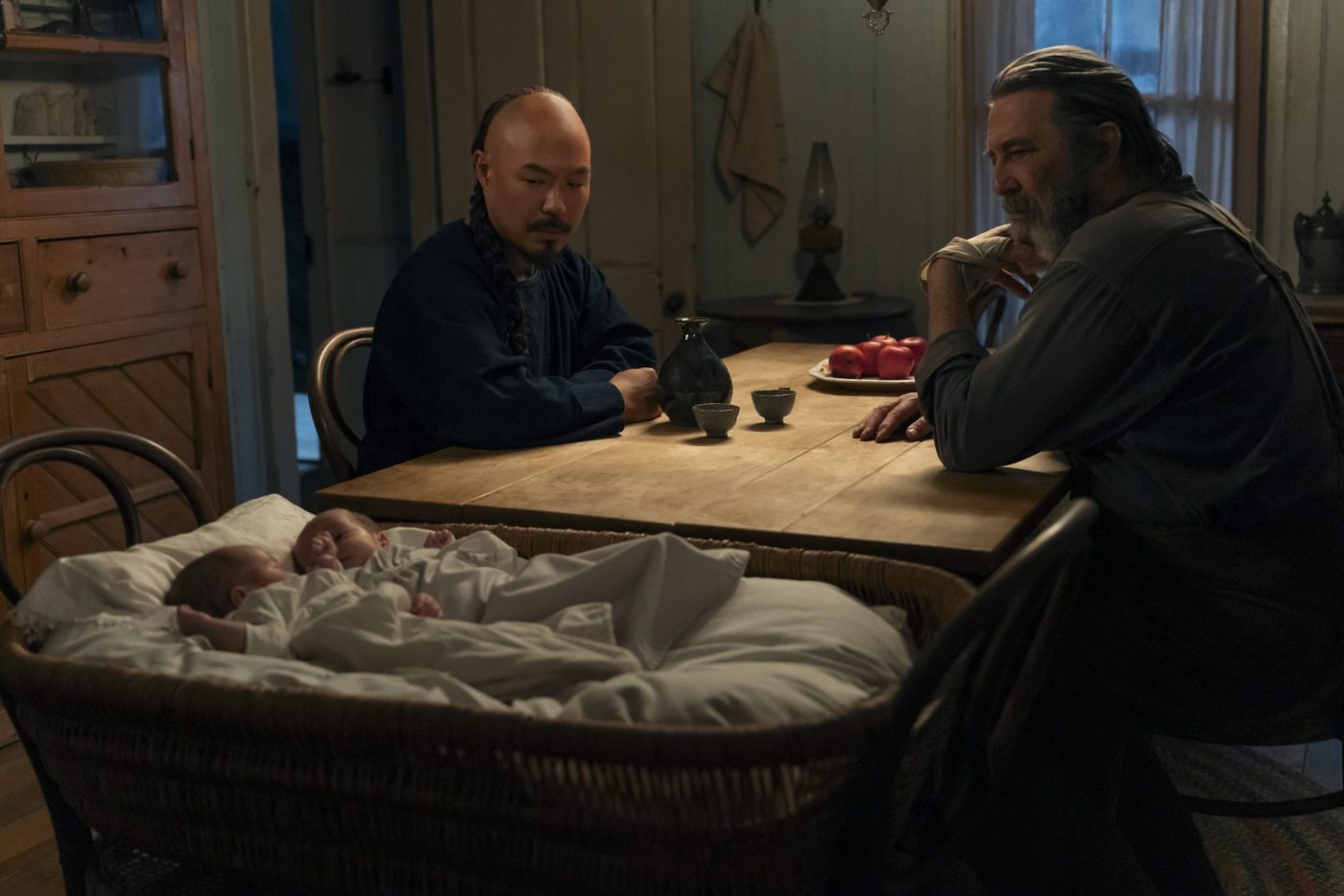 "Two men sit at a wooden table in a dimly lit rustic room, with a basket containing two sleeping babies placed in front of them; shelves and kitchen items are visible in the background."