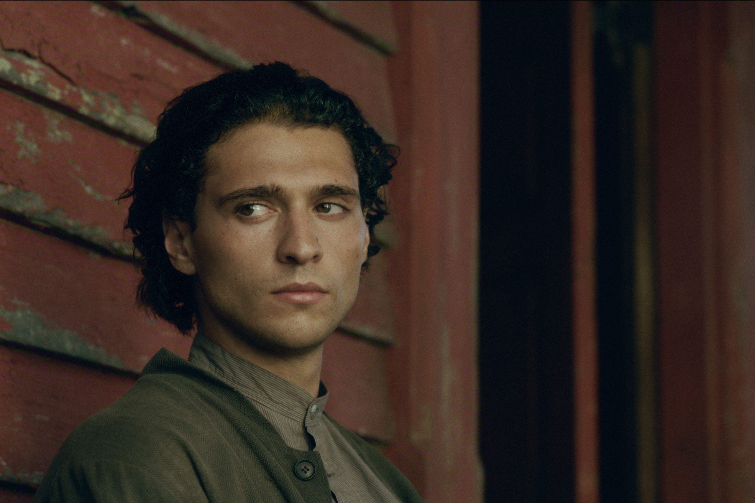 "Young man with dark hair wearing a buttoned shirt and jacket, standing in front of weathered red wooden wall, looking to the side with a thoughtful expression in a rustic, moody outdoor setting."