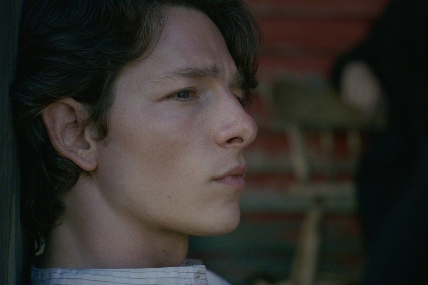 "Young man with wavy hair sits indoors, close to a wooden wall and an old wooden chair, appearing thoughtful and gazing off to the side in soft, natural light."